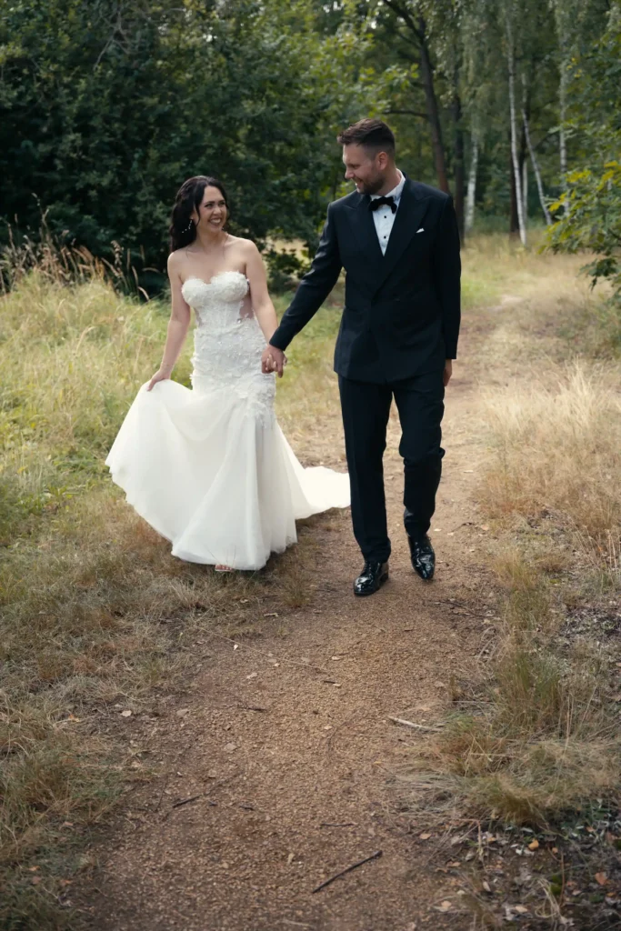Brautpaar geht lächelnd Hand in Hand über einen Weg zwischen Bäumen und Gräsern – Paarmotiv zur Hochzeit in Bautzen.