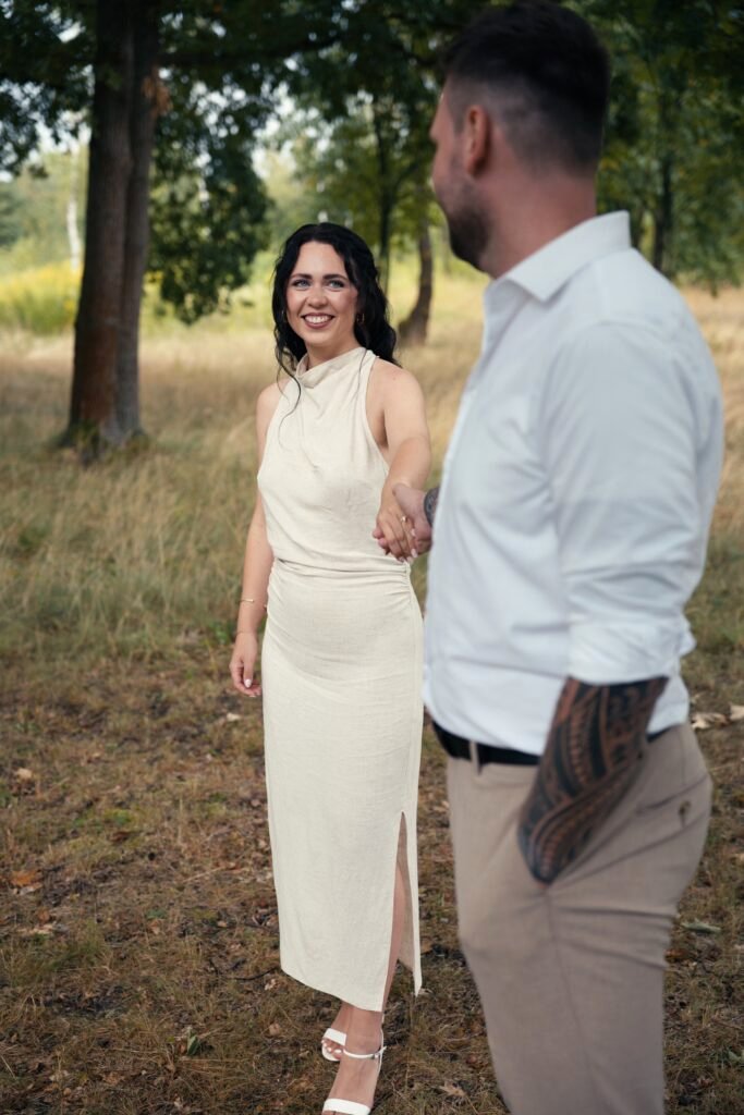 Paar in heller Kleidung geht Hand in Hand zwischen Bäumen und schaut sich an – Paarmotiv zur Hochzeit in Dresden.