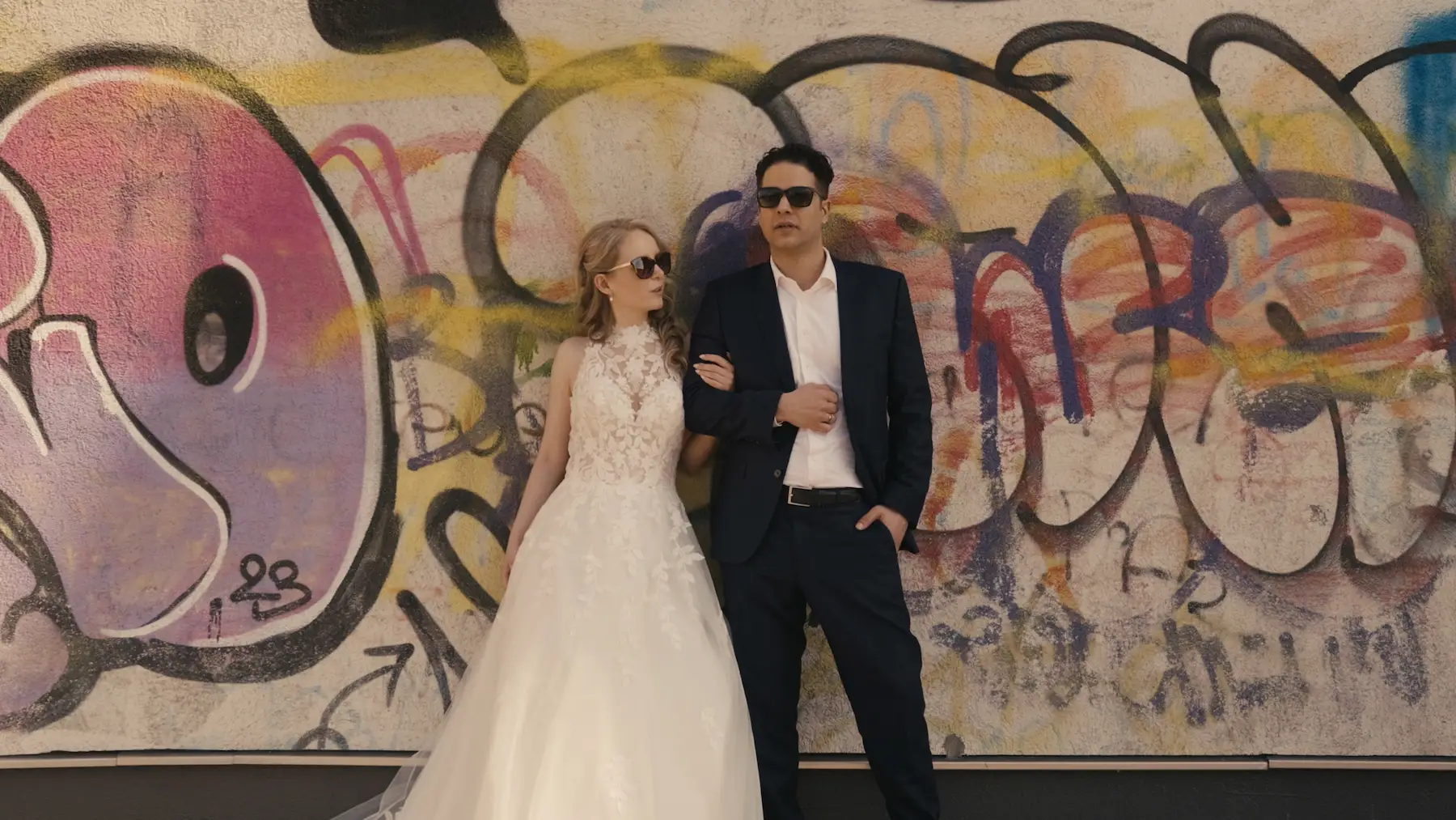 Brautpaar mit Sonnenbrillen steht Arm in Arm vor einer bunten Graffitiwand – Hochzeitsfilm in Dresden.