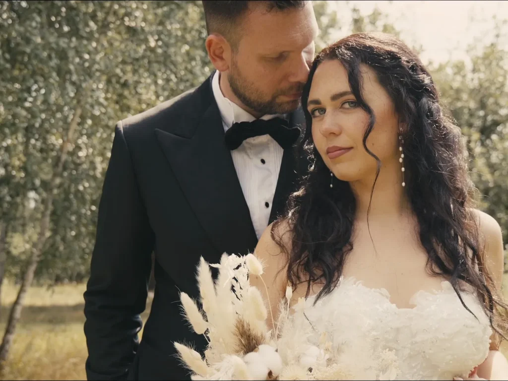 Braut mit Trockenblumenstrauß und Bräutigam im schwarzen Anzug stehen eng beieinander unter Bäumen – Hochzeitsfilm in Dresden.