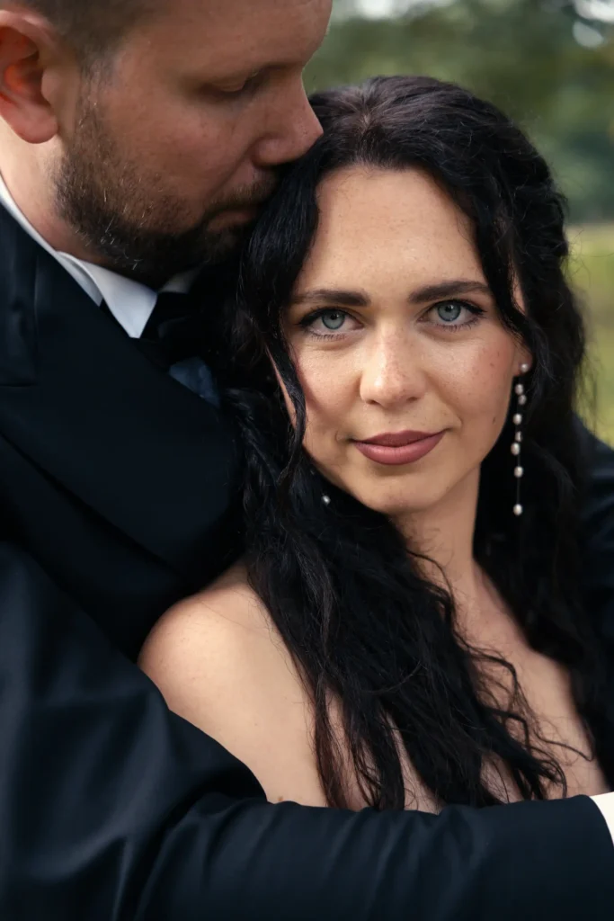Hochzeitspaar in enger Umarmung: Der Bräutigam küsst die Braut auf den Kopf, während sie in die Kamera blickt.