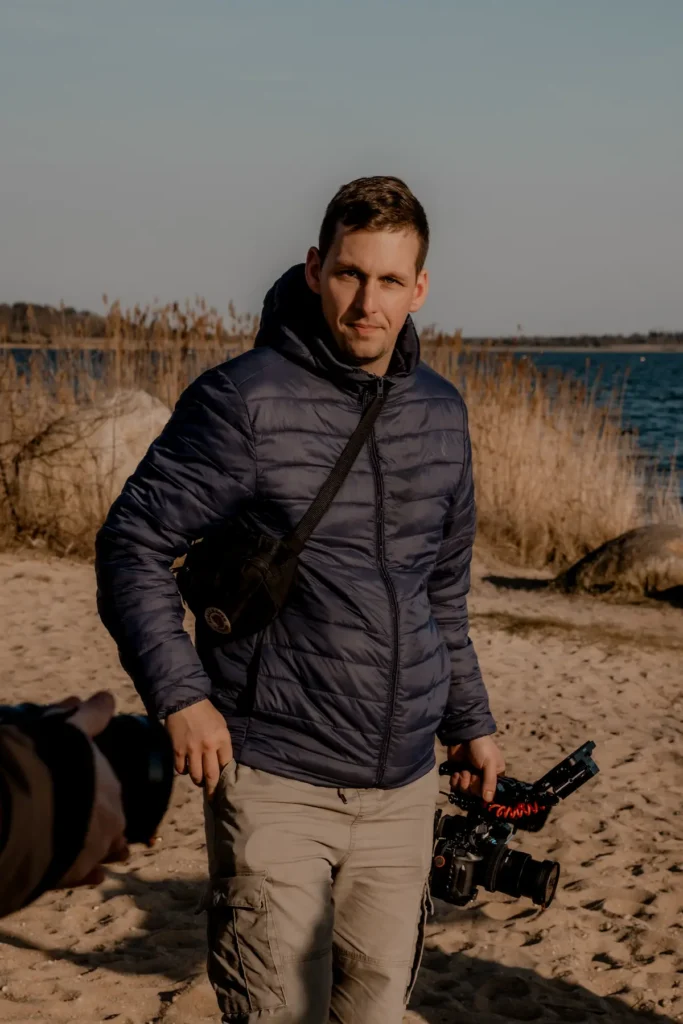 Hochzeitsvideograf am Strand mit Filmkamera in der Hand, dunkler Steppjacke und Schilf im Hintergrund.