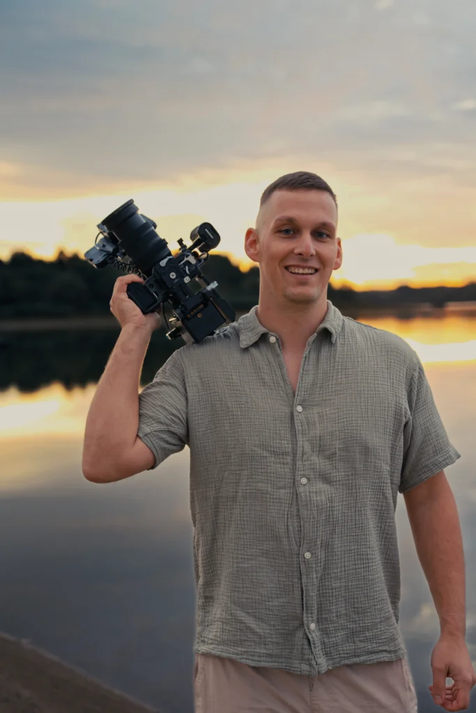 Hochzeitsvideograf Stefan Bergner aus Bautzen lächelt mit einer Filmkamera auf der Schulter am See im Sonnenuntergang.